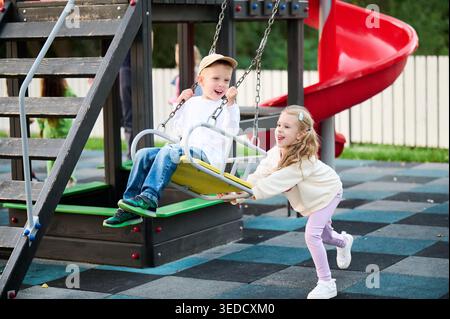 Girl pushing boy on swing, both laughing and enjoying fast movement on playground, active kids outdoor play. Concept of friendship, trust, energy, carefree childhood Stock Photo