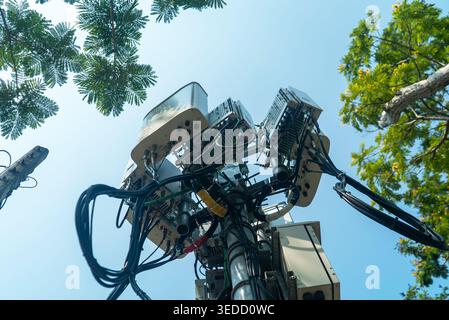 A view from below of a communication tower surrounded by greenery, showcasing antennas and equipment against a clear blue sky. Ideal for technology th Stock Photo