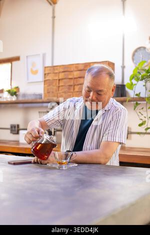 Senior Asian man pouring tea from glass teapot into cup at counter with smartphone, striped shirt Stock Photo