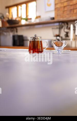 Glass teapot is steeping tea on pale countertop with empty teacup and sunlit window, copy space Stock Photo