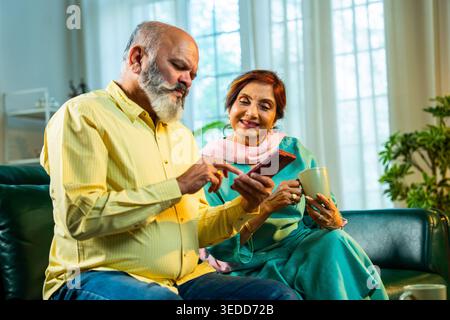 Retired senior Indian Asian old age couple using a smartphone together while sitting on a sofa at home in cozy interiors; lifestyle, digital technolog Stock Photo