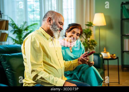 Retired senior Indian Asian old age couple using a smartphone together while sitting on a sofa at home in cozy interiors; lifestyle, digital technolog Stock Photo