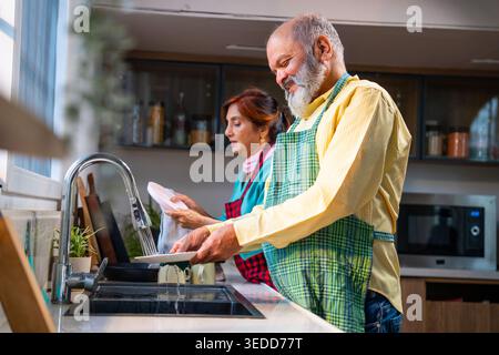 Retired senior Indian Asian old age couple together washing dishes at a modern kitchen platform; showcasing teamwork, daily household chores, and toge Stock Photo