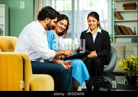 Indian young happy couple discussing financial or legal matters with female corporate consultant during home meeting while reviewing documents and pla Stock Photo
