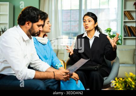 Indian young happy couple discussing financial or legal matters with female corporate consultant during home meeting while reviewing documents and pla Stock Photo