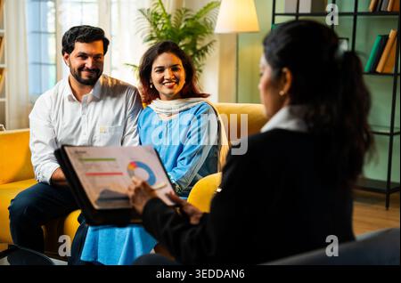 Indian young happy couple discussing financial or legal matters with female corporate consultant during home meeting while reviewing documents and pla Stock Photo