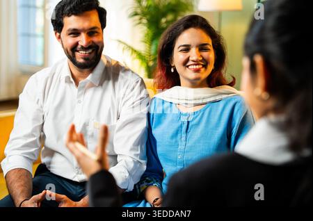Closeup of Indian young happy couple discussing with female corporate consultant during home meeting while smiling and reviewing financial documents f Stock Photo