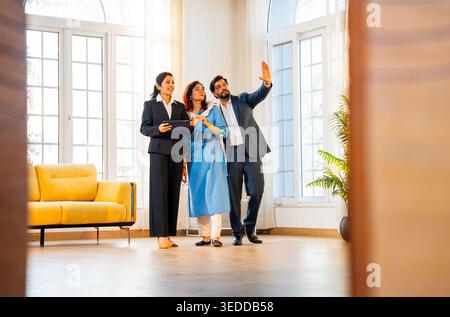 Indian woman real estate agent with young married couple walking in new property with tablet to discuss interior layout and design changes during a pr Stock Photo
