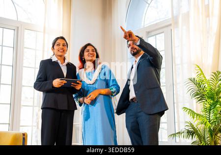 Indian woman real estate agent with young married couple walking in new property with tablet to discuss interior layout and design changes during a pr Stock Photo