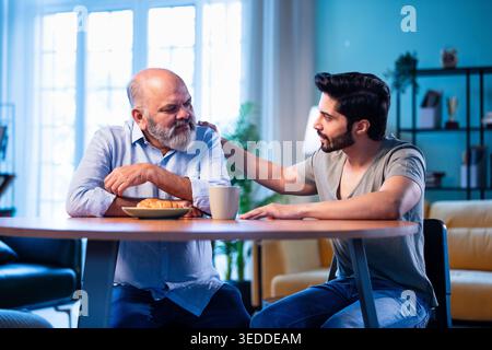 Indian young son having serious conversation with senior father in modern living room at home, emotional family discussion between two generations sha Stock Photo