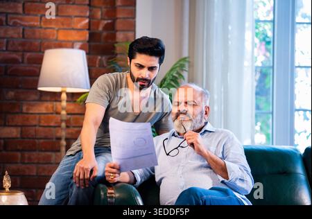 Indian retired father and young son discussing finance while reviewing financial report, profit and loss statement and business growth documents toget Stock Photo