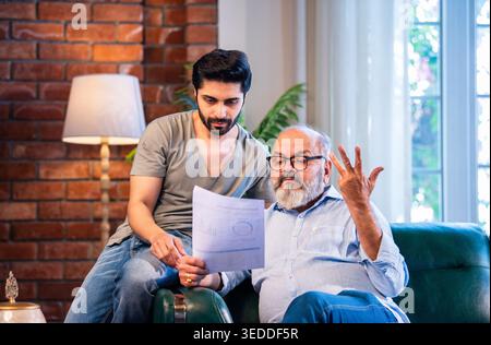 Indian retired father and young son discussing finance while reviewing financial report, profit and loss statement and business growth documents toget Stock Photo