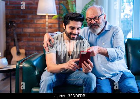 Indian young son and senior father using smartphone together while sitting on sofa in modern home, browsing internet, watching funny reels and photos, Stock Photo