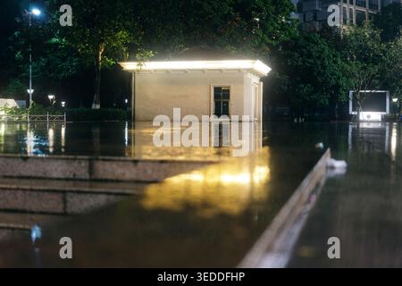 Nighttime urban flood scene with illuminated pavilion reflecting in water, wet pavement, and city Stock Photo