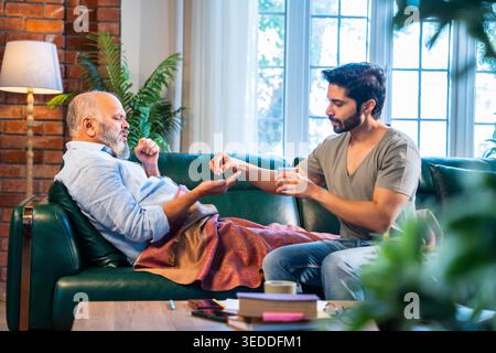 Indian retired father in poor health resting on sofa at home while young son giving medicine and glass of water, caring family moment showing support, Stock Photo
