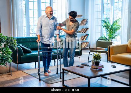 Indian young son helping and assisting aged father stand and walk with his physical support in living room at modern home, showing patience, strength Stock Photo
