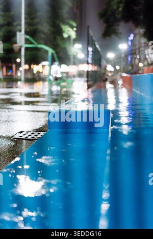 Nighttime urban pool edge with reflective blue water, wet pavement, and blurred city lights in Stock Photo