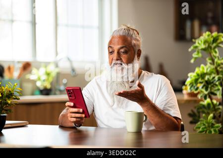 Indian mid age stylish man with long white beard and hair using smartphone while sitting at breakfast table in modern kitchen interior, browsing inter Stock Photo