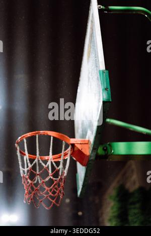 Close-up of basketball hoop with orange rim and worn net, mounted on green backboard against dark Stock Photo