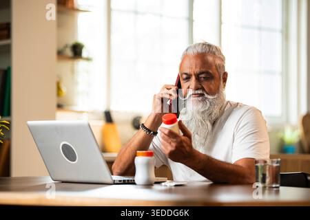 Indian mid age man checking medicine ingredients online holding pills bottle while speaking on phone and using laptop at table in modern kitchen inter Stock Photo