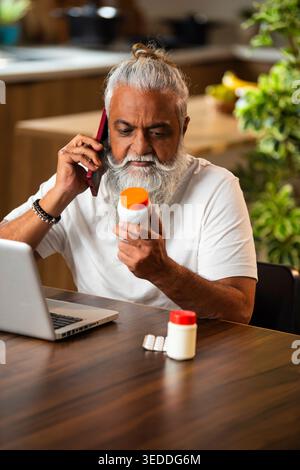 Indian mid age man checking medicine ingredients online holding pills bottle while speaking on phone and using laptop at table in modern kitchen inter Stock Photo