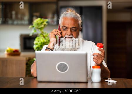 Indian mid age man checking medicine ingredients online holding pills bottle while speaking on phone and using laptop at table in modern kitchen inter Stock Photo