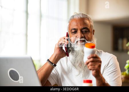 Indian mid age man checking medicine ingredients online holding pills bottle while speaking on phone and using laptop at table in modern kitchen inter Stock Photo