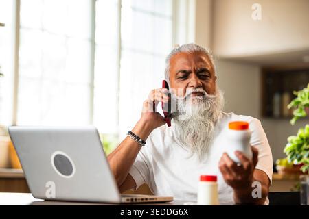 Indian mid age man checking medicine ingredients online holding pills bottle while speaking on phone and using laptop at table in modern kitchen inter Stock Photo