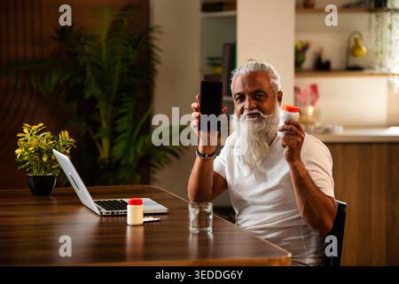 Indian mid age man checking medicine ingredients online holding pills bottle while speaking on phone and using laptop at table in modern kitchen inter Stock Photo