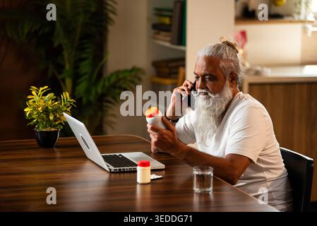 Indian mid age man checking medicine ingredients online holding pills bottle while speaking on phone and using laptop at table in modern kitchen inter Stock Photo