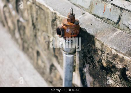 Rusty fire hydrant mounted on weathered stone wall with pipe, close-up urban infrastructure detail Stock Photo