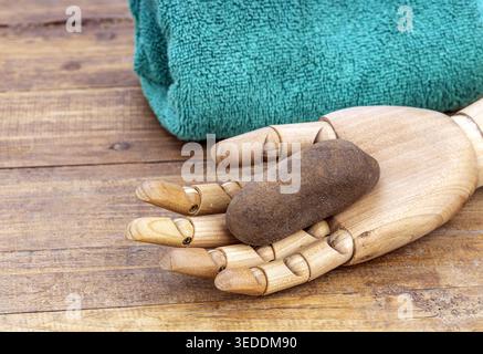 Close-up of a jointed wooden artist mannequin hand cradling a rough dark wet pumice stone to exfoliate dry skin, resting on weathered wood planks next Stock Photo