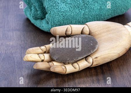 A detailed articulated wooden mannequin hand gently holds a wet smooth oval gray pumice stone to exfoliate rough skin, with a soft teal green towel re Stock Photo
