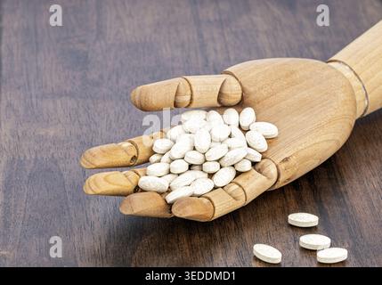 An articulated wooden artist mannequin hand cradles a large pile of white oval exfoliate supplement tablets. Several pills are scattered on the dark w Stock Photo