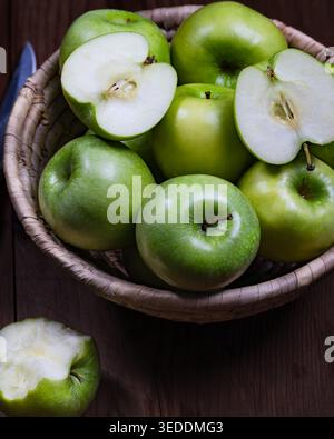 A close-up view of fresh green apples in a woven basket on a dark wooden surface, with two apple halves showing the juicy interior. The image highligh Stock Photo