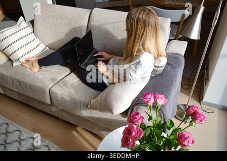 Woman working on a laptop on a comfortable sofa in a stylish apartment with pink roses in the foreground Stock Photo