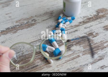 Close-up of a hand holding glasses pointed at a trail of white and blue pills leading to a pharmacy bottle on an old wooden table, for treatment strat Stock Photo