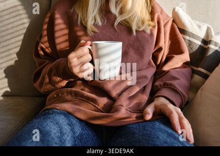 Blonde woman's hands gently holding a white ceramic mug with a hot drink while wearing a cozy brown sweater in soft morning light Stock Photo