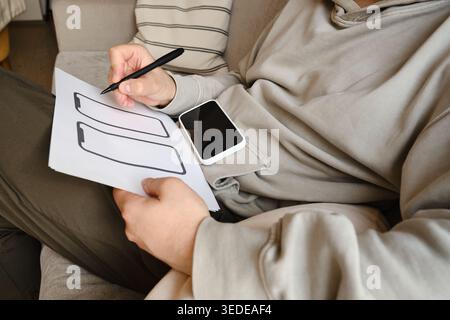 Person holding a smartphone and pointing at a paper wireframe template on a white round table Stock Photo