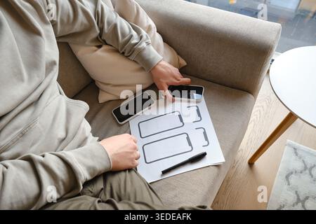 Person holding a smartphone and pointing at a paper wireframe template on a white round table Stock Photo