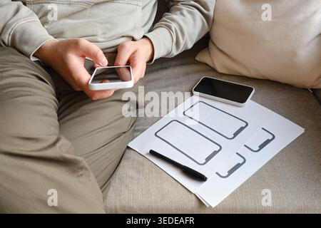 Person holding a smartphone and pointing at a paper wireframe template on a white round table Stock Photo