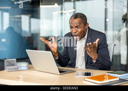 Angry businessman expressing frustration and stress while looking at a laptop screen in a modern office, reacting to a professional error or conflict with raised hands Stock Photo
