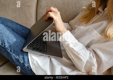 Woman in a white shirt and jeans sitting on a sofa and spreading her hands in confusion while looking at a laptop Stock Photo