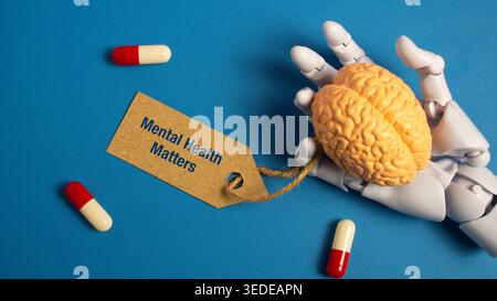 Robotic hand holding a model of a human brain with a tag saying mental health matters and pills on a blue background for AI therapy concepts. Stock Photo