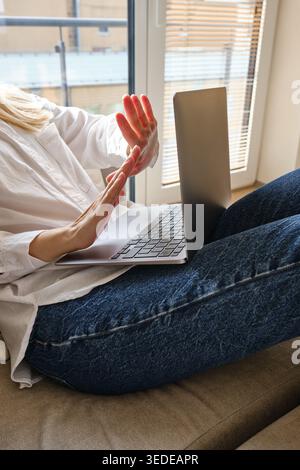 Woman in a white shirt and jeans sitting on a sofa and spreading her hands in confusion while looking at a laptop Stock Photo