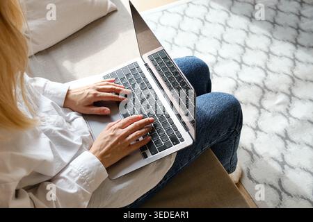 Woman in a white shirt and jeans sitting on a sofa and spreading her hands in confusion while looking at a laptop Stock Photo