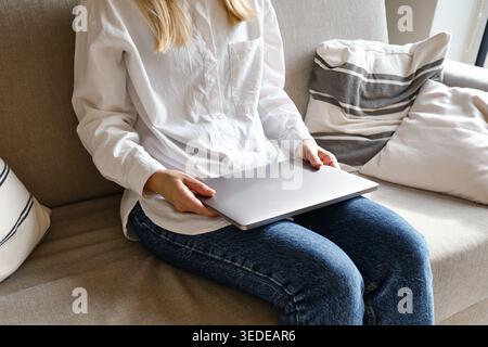 Woman in a white shirt and jeans sitting on a sofa and holding a closed laptop on her lap Stock Photo