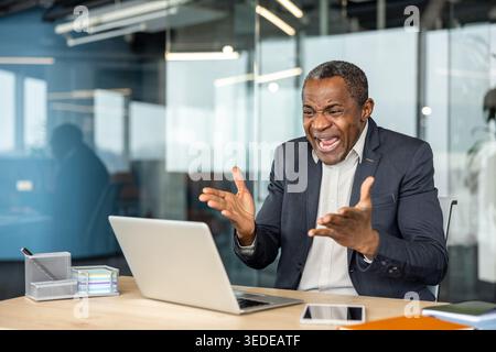 Stressed mature professional man throwing hands up in exasperation and shouting at his laptop, experiencing a computer problem or work pressure during an intense business day in the office Stock Photo