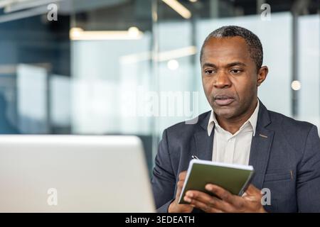 Focused businessman is taking notes in a notebook while looking at a laptop screen, engaging in a concentrated virtual meeting or online learning session in a modern office setup Stock Photo