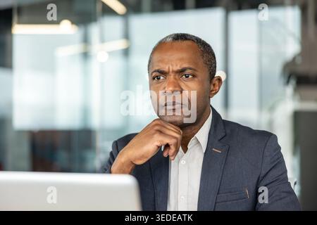 Mature man with a serious expression, hand on chin, thinking deeply while working on a laptop in a modern office, representing concentration and problem-solving Stock Photo